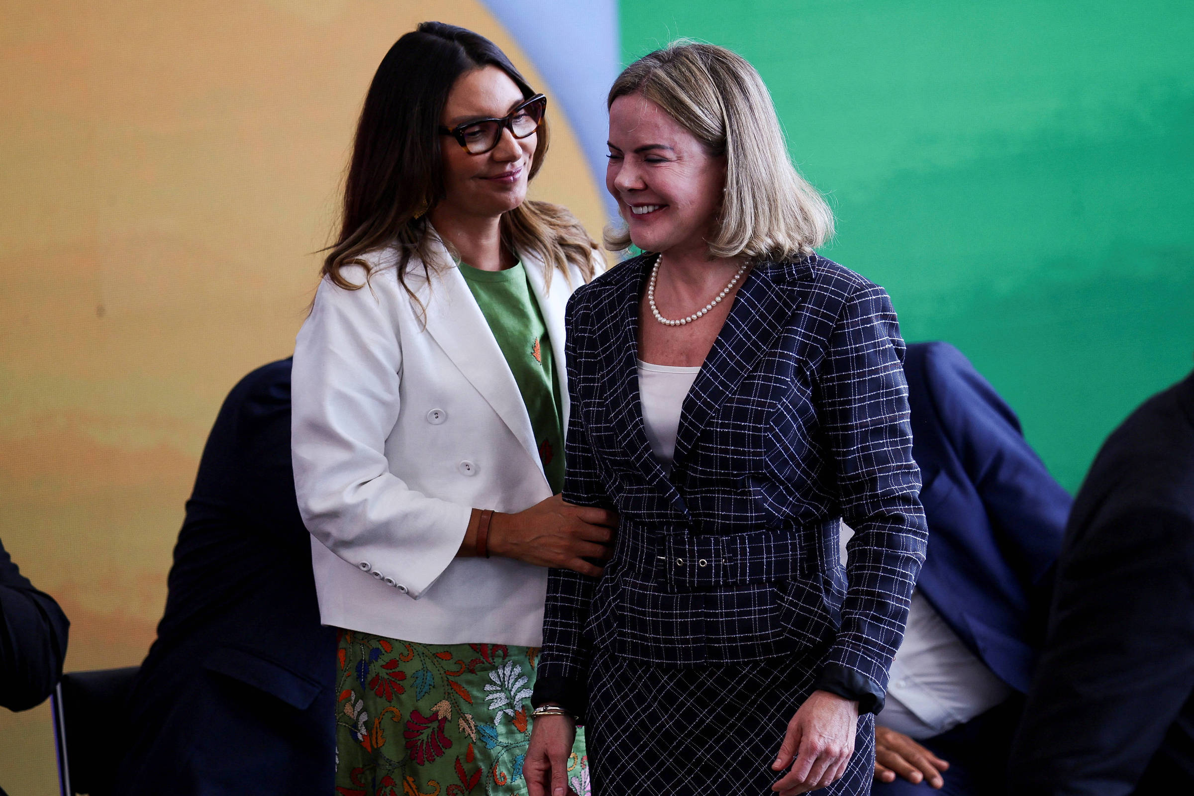 A imagem mostra duas mulheres em um evento. A mulher à esquerda usa óculos, uma blusa verde e um casaco branco, com uma saia estampada. A mulher à direita está vestindo um vestido xadrez e parece sorrir enquanto a outra mulher a toca levemente nas costas. Ao fundo, há uma parede com cores em degradê.