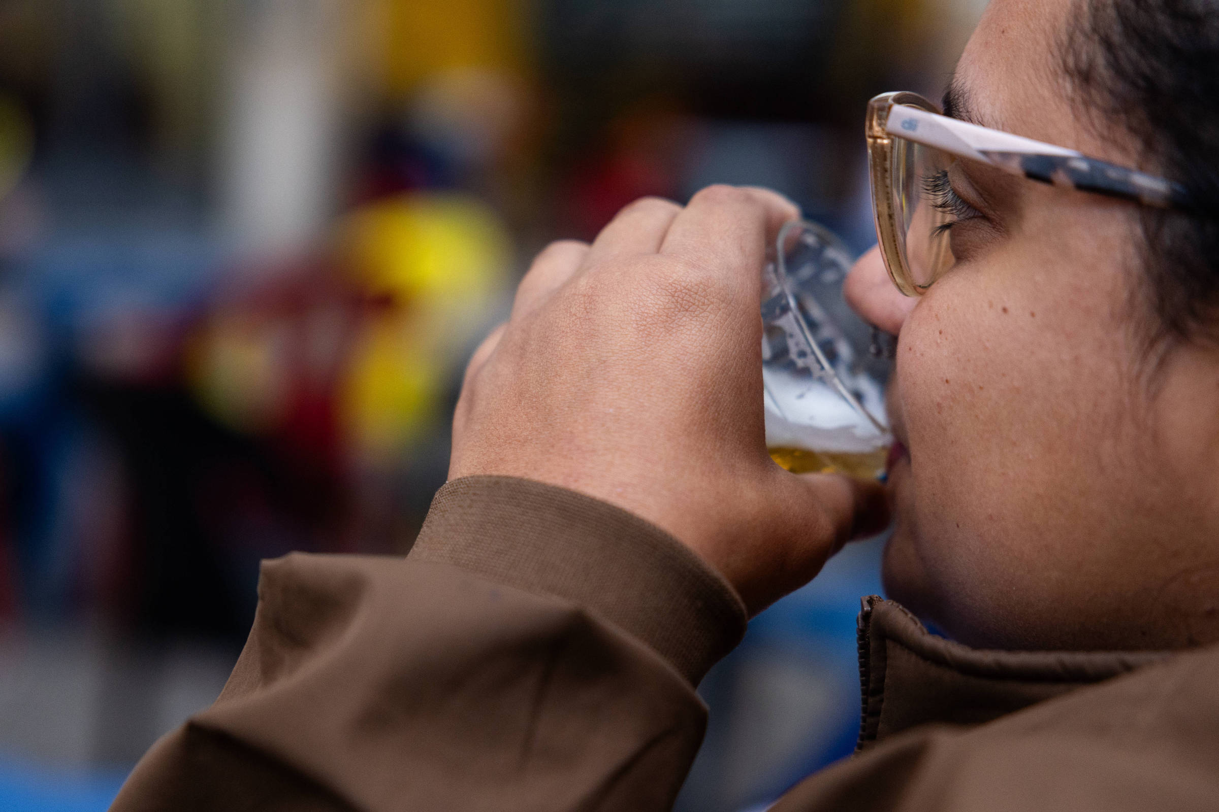 Pessoa de perfil usando óculos transparentes e casaco marrom segura e bebe de copo plástico com cerveja, com fundo desfocado de ambiente externo.