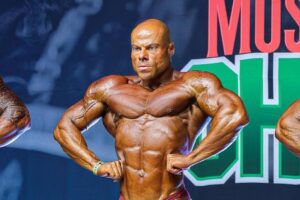 Fisiculturista masculino no centro do palco em pose de frente, exibindo musculatura definida e bronzeada, usando sunga vermelha. Dois outros competidores parcialmente visíveis nas laterais. Fundo com banners de patrocinadores e iluminação de palco.