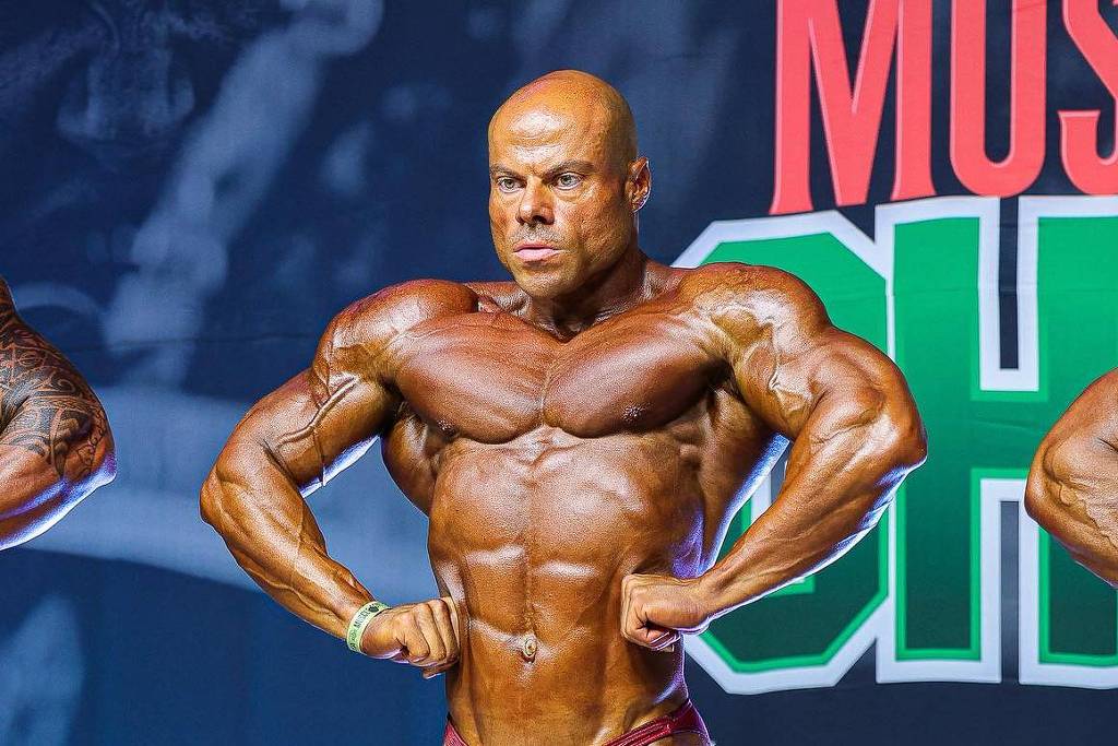 Fisiculturista masculino no centro do palco em pose de frente, exibindo musculatura definida e bronzeada, usando sunga vermelha. Dois outros competidores parcialmente visíveis nas laterais. Fundo com banners de patrocinadores e iluminação de palco.