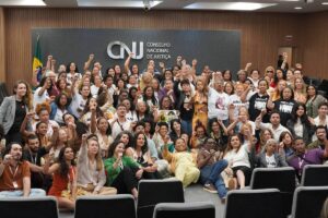 Cerca de 80 pessoas reunidas em auditório do Conselho Nacional de Justiça, muitas com punhos erguidos. Ambiente interno com parede de madeira e bandeira do Brasil ao fundo.