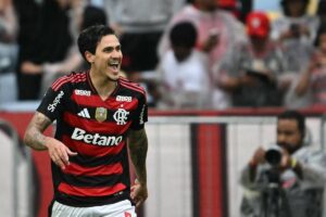 Jogador do Flamengo com uniforme vermelho e preto sorri enquanto comemora em campo. Torcedores e fotógrafos aparecem desfocados ao fundo.