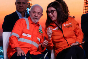 A imagem mostra duas pessoas sentadas em um evento. À esquerda, um homem idoso com cabelo grisalho, usando uma camisa laranja com detalhes cinzas e uma bandeira do Brasil no peito. À direita, uma mulher com cabelo longo e escuro, vestindo uma jaqueta laranja com o logotipo da Petrobras. Ambos parecem estar conversando e sorrindo, com um fundo desfocado de um cenário urbano ao pôr do sol.