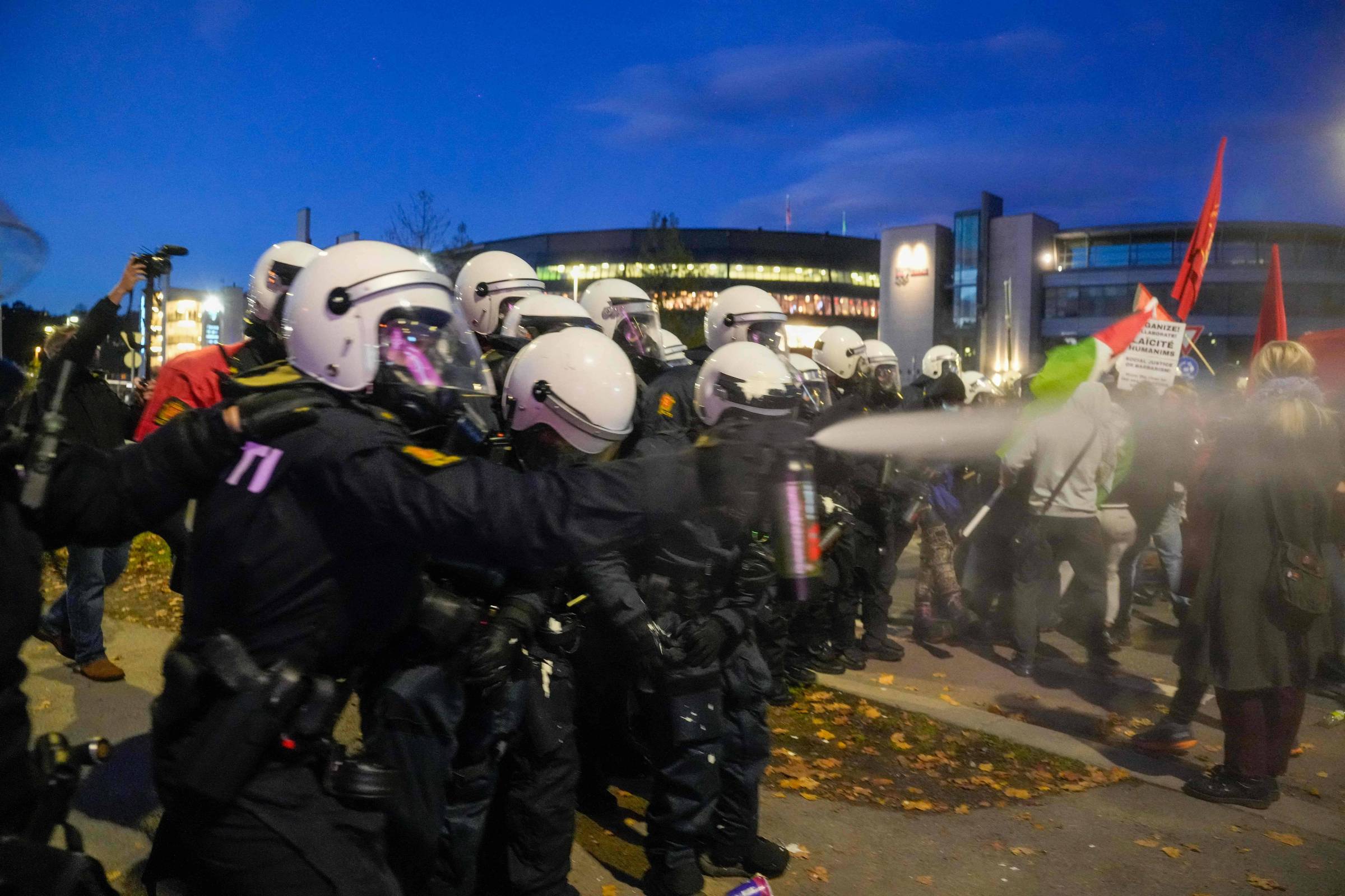 Policiais em linha, usando capacetes brancos e uniformes pretos, dispersam manifestantes com spray de pimenta em área externa urbana ao anoitecer. Manifestantes seguram bandeiras vermelhas e cartazes.