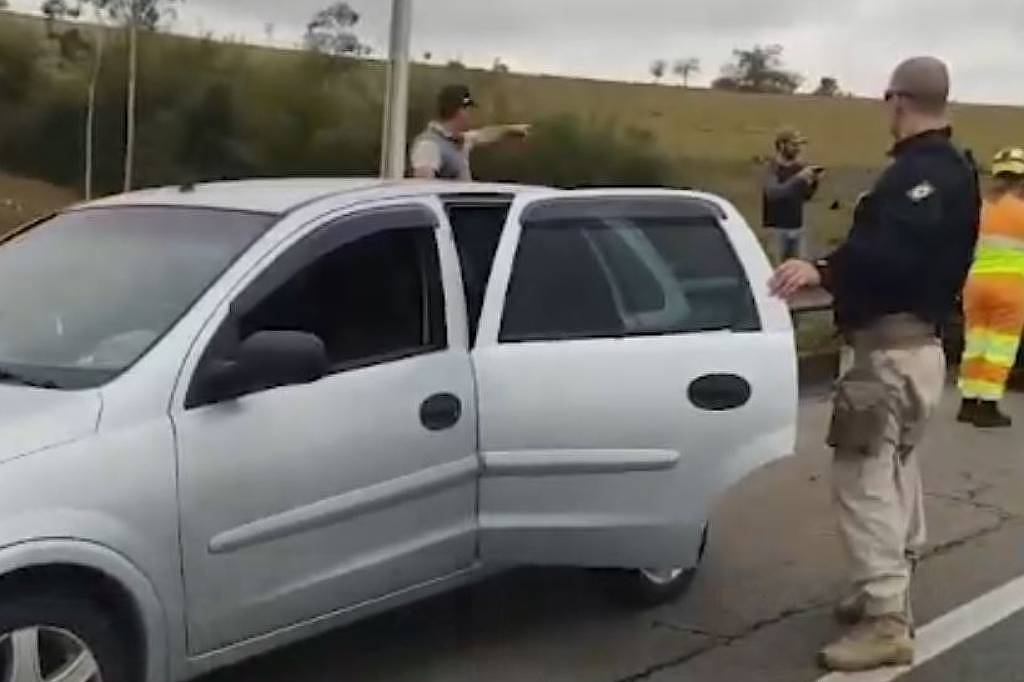 Policial em uniforme e capacete orienta condutor de carro prata com porta aberta em estrada. Ao fundo, homem aponta para frente e trabalhador com roupa laranja e amarela. Céu nublado e vegetação ao redor.