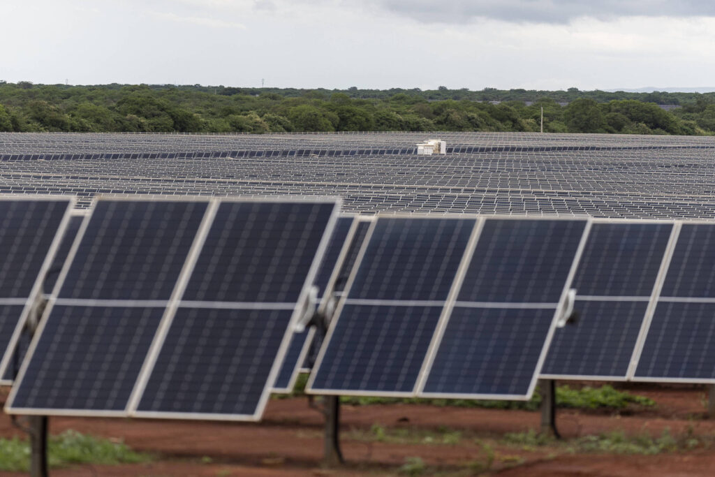 A imagem mostra um grande campo de painéis solares dispostos em fileiras. Os painéis são predominantemente de cor escura e estão posicionados em um terreno de terra vermelha. Ao fundo, há uma vasta área verde com árvores e um céu nublado.