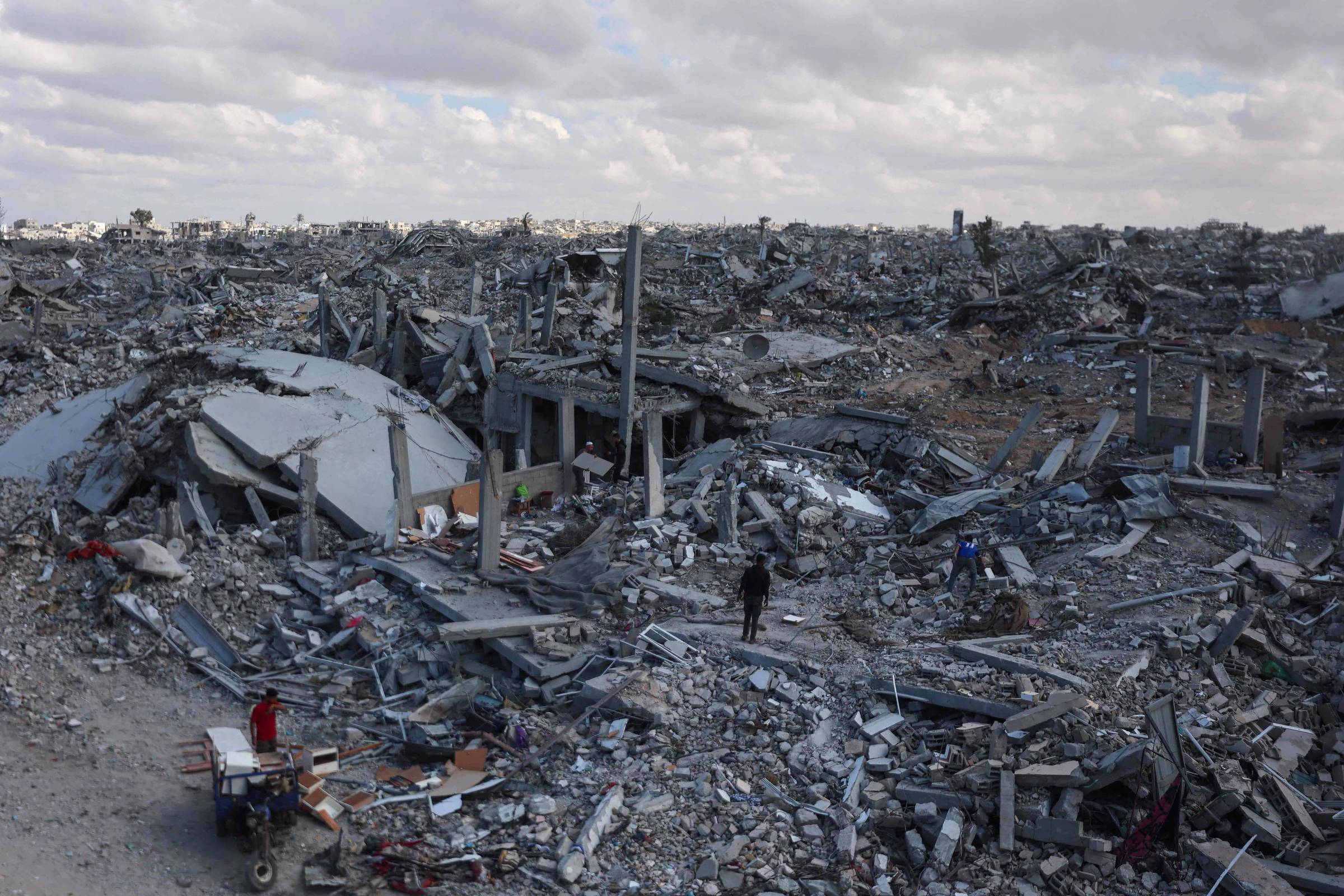 Grande extensão de destroços de edifícios destruídos sob céu nublado. Três pessoas caminham entre os escombros, destacando a escala da destruição.