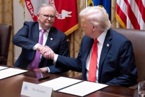 Dois homens sentados à mesa apertam as mãos. Um deles usa terno escuro com gravata vermelha e tem cabelo branco; o outro usa terno escuro com gravata lilás e óculos. Bandeiras oficiais estão ao fundo.