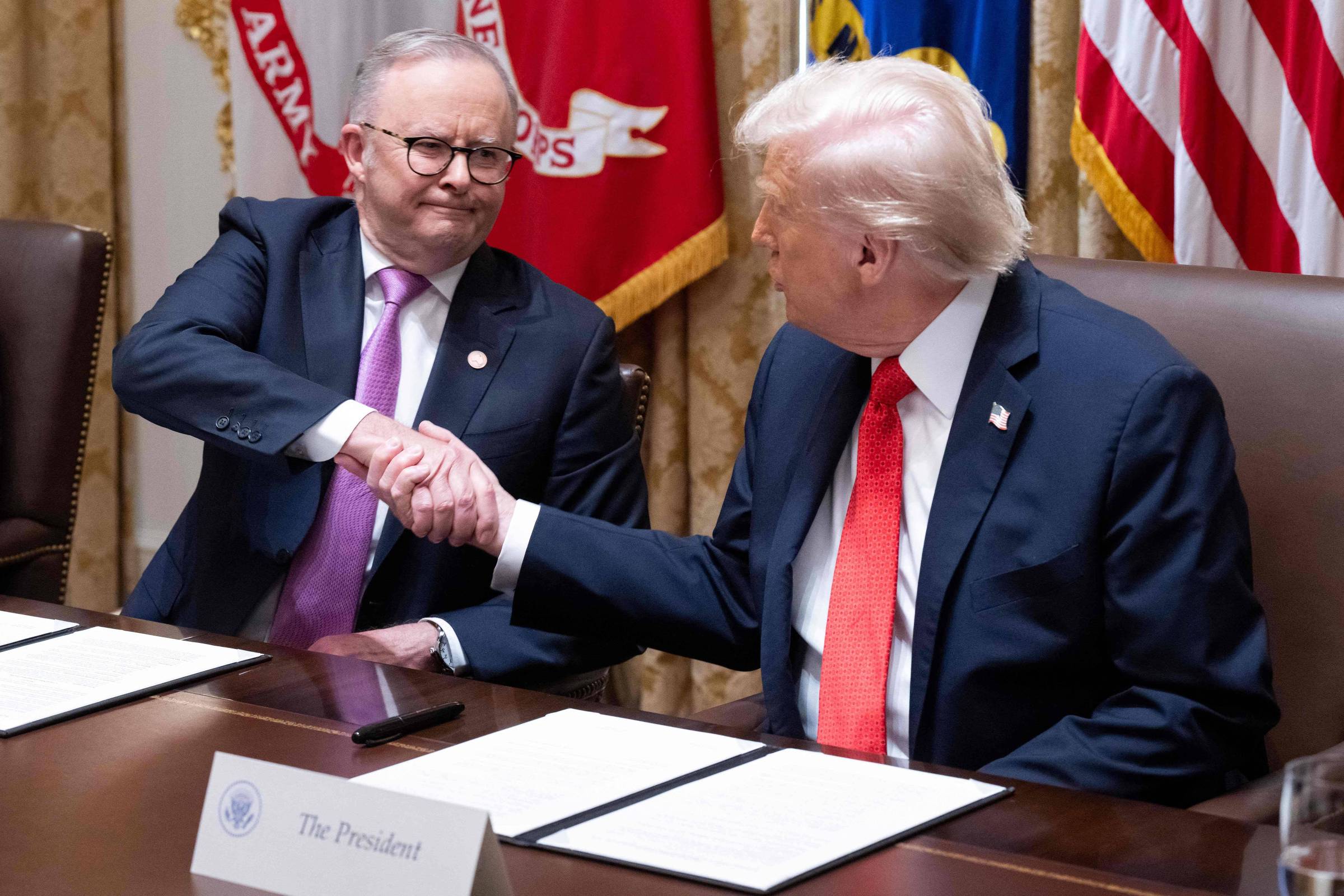 Dois homens sentados à mesa apertam as mãos. Um deles usa terno escuro com gravata vermelha e tem cabelo branco; o outro usa terno escuro com gravata lilás e óculos. Bandeiras oficiais estão ao fundo.