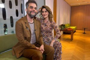 Homem e mulher sentados lado a lado em sofá verde em sala com piso de madeira e paredes claras. Homem veste terno marrom e camiseta cinza, mulher usa vestido estampado com padrão geométrico. Ao fundo, cortinas brancas, mesa de madeira com vaso de plantas e poltrona verde.