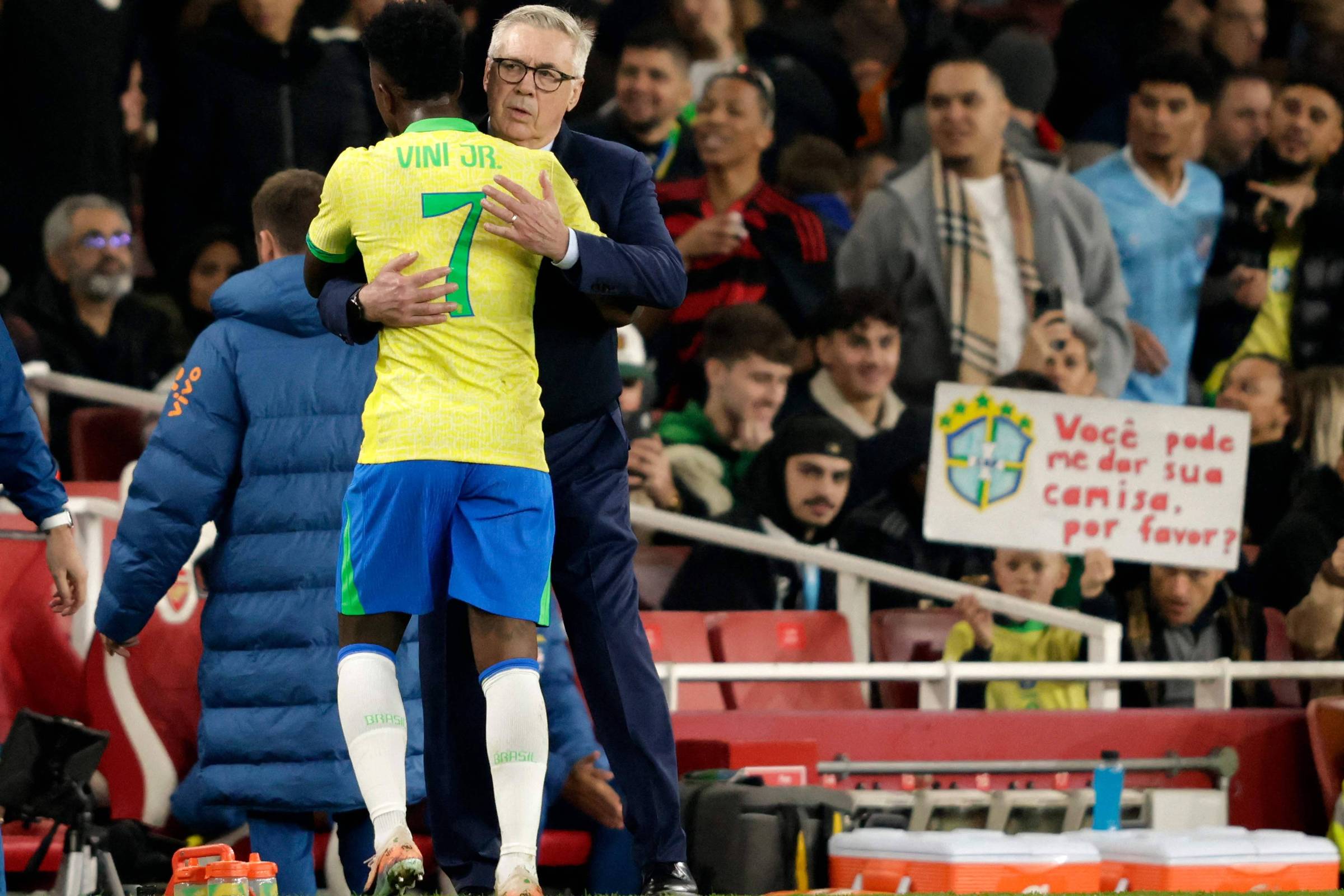 Jogador com uniforme amarelo e azul, número 7, abraça treinador de terno escuro na beira do campo. Torcida ao fundo segura placa pedindo camisa do jogador.
