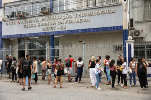 Grupo de pessoas em fila do lado de fora do Centro Federal de Educação Tecnológica Celso Suckow da Fonseca, em dia claro. Fachada do prédio com letreiro em destaque e portão de grades fechado.