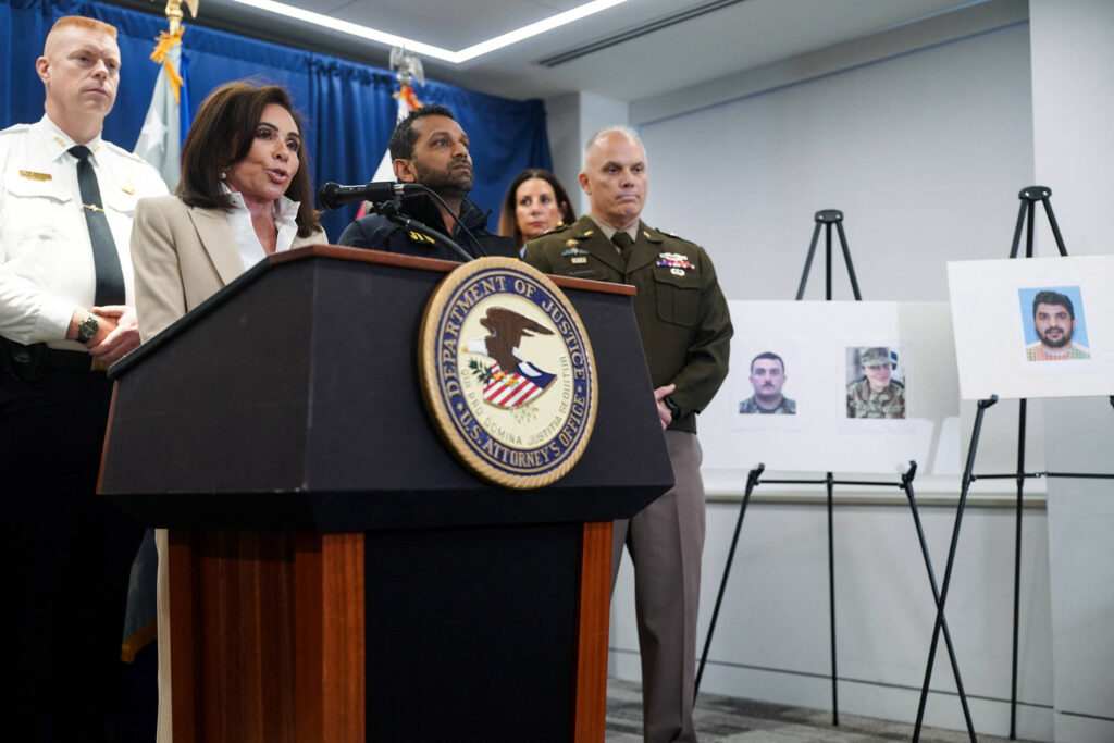 Cinco autoridades, incluindo policiais e militares, participam de coletiva em sala oficial. Mulher fala no púlpito com selo do Departamento de Justiça dos EUA. Ao lado, três retratos em cavaletes exibem fotos de homens.
