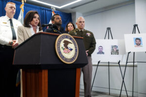 Cinco autoridades, incluindo policiais e militares, participam de coletiva em sala oficial. Mulher fala no púlpito com selo do Departamento de Justiça dos EUA. Ao lado, três retratos em cavaletes exibem fotos de homens.