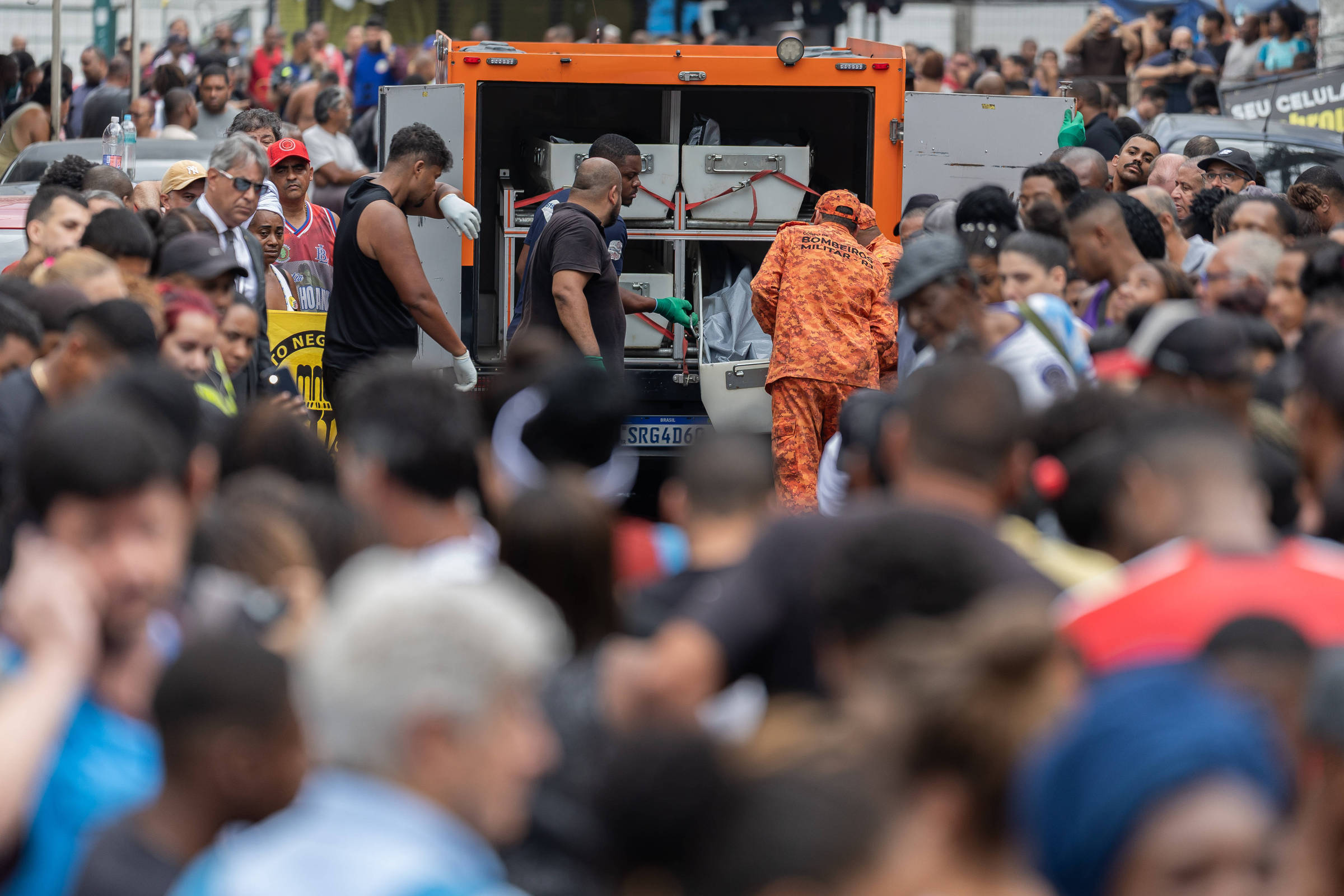 Equipe de resgate com uniforme laranja e preto carrega corpo coberto em maca para caminhão de resgate aberto. Multidão observa ao redor em ambiente urbano durante o dia.