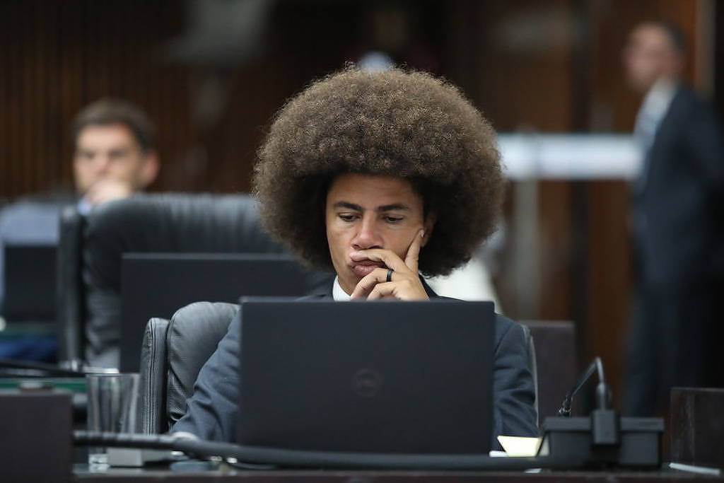 Um jovem com cabelo afro volumoso está sentado em uma mesa de trabalho, olhando para um laptop. Ele está vestido com um terno escuro e uma camisa clara, com uma expressão pensativa. Ao fundo, há outras pessoas e mesas, sugerindo um ambiente de trabalho ou reunião.
