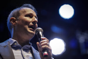 Um homem está falando ao microfone em um palco. Ele usa um paletó cinza e uma camisa clara. O fundo é escuro, com luzes brilhantes em destaque, criando um ambiente de apresentação ou evento. O homem parece estar em um momento de discurso ou apresentação.