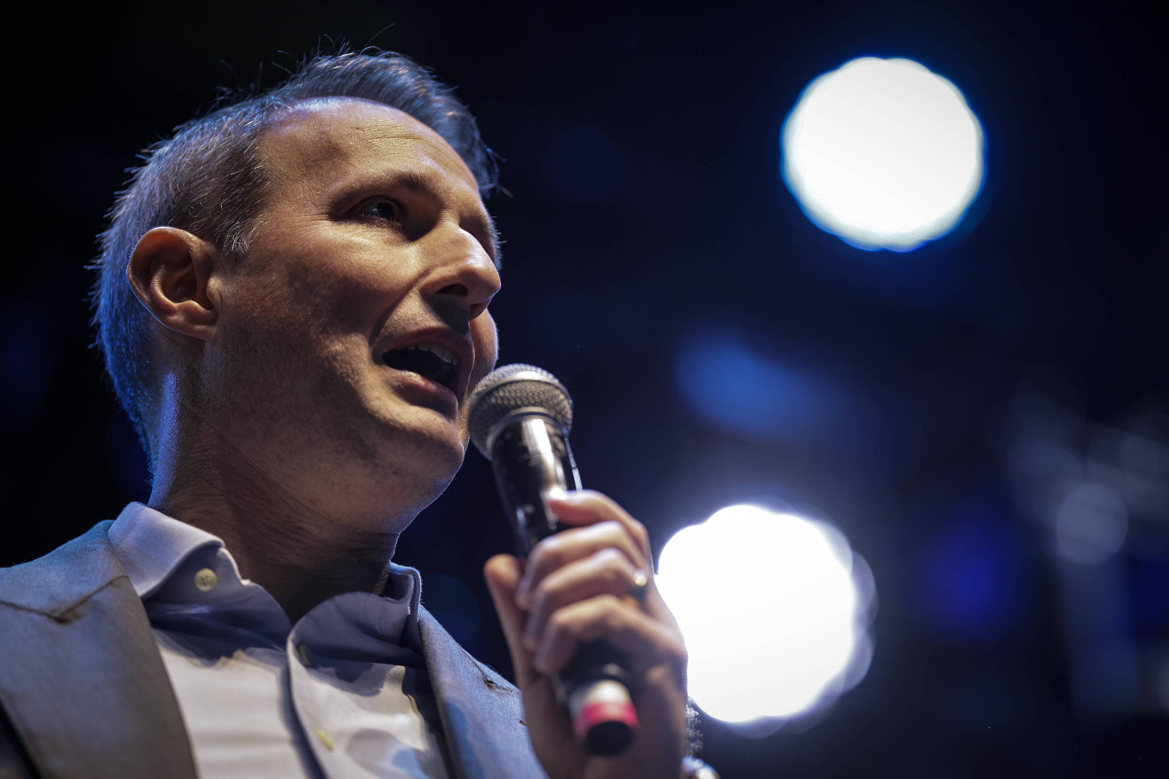 Um homem está falando ao microfone em um palco. Ele usa um paletó cinza e uma camisa clara. O fundo é escuro, com luzes brilhantes em destaque, criando um ambiente de apresentação ou evento. O homem parece estar em um momento de discurso ou apresentação.