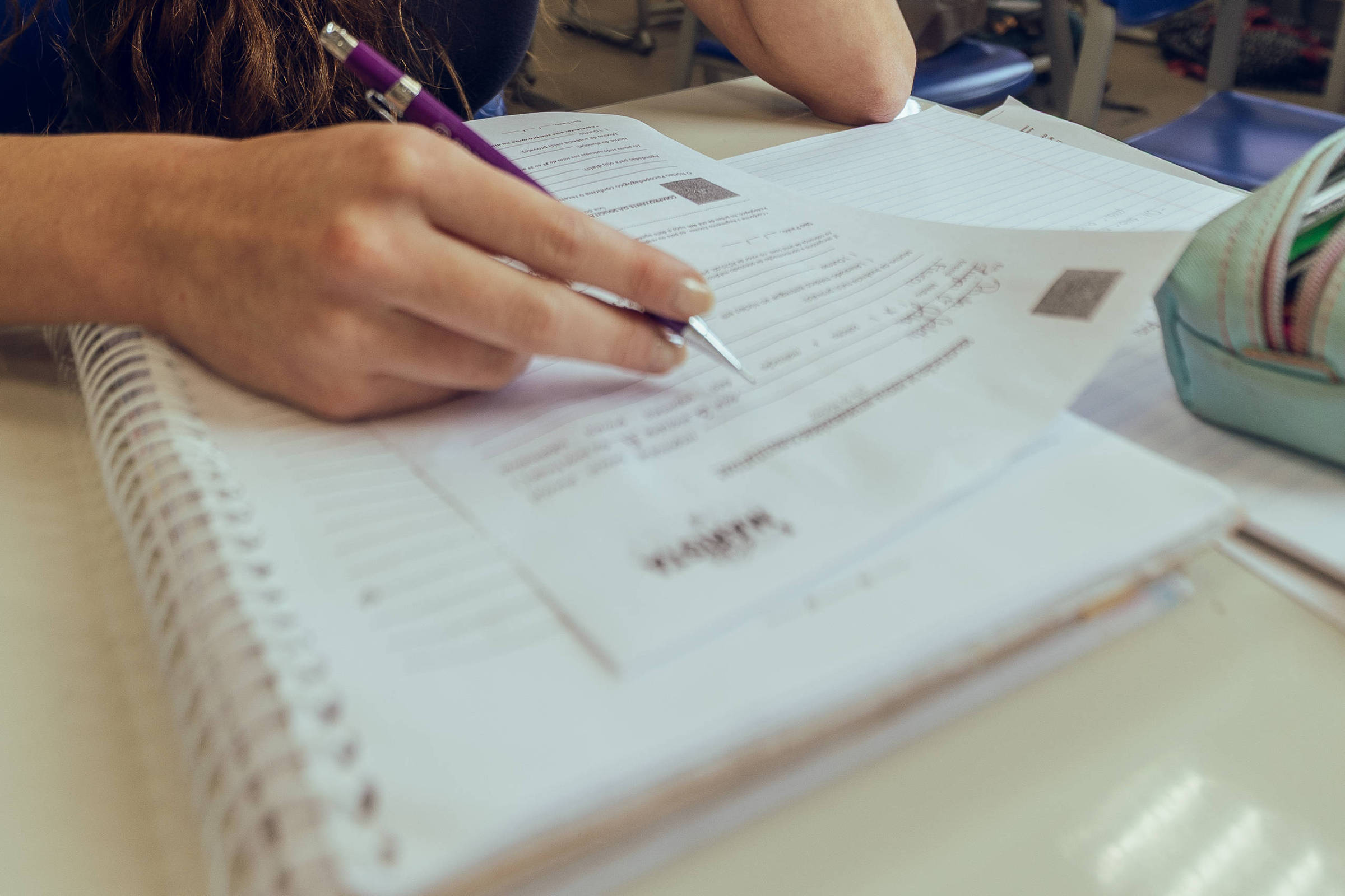 Jovem sentada em carteira de sala de aula iluminada, escrevendo em caderno espiral com caneta roxa. Ao fundo, carteiras vazias e janelas com persianas parcialmente fechadas.