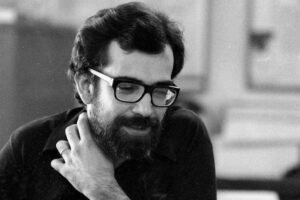 A imagem mostra um homem com cabelo escuro e barba, usando óculos. Ele está vestindo uma camisa preta e parece estar pensativo, com a mão no pescoço. O fundo é desfocado, sugerindo um ambiente interno.