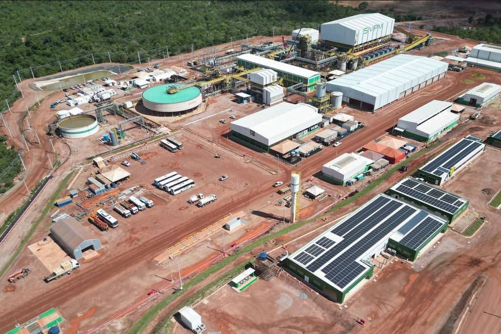 A imagem mostra uma vista aérea de uma instalação industrial localizada em uma área com vegetação ao fundo. A estrutura inclui vários edifícios, tanques e áreas de armazenamento. Há também painéis solares visíveis em alguns dos telhados. O solo é predominantemente de terra batida, e há veículos estacionados nas proximidades.
