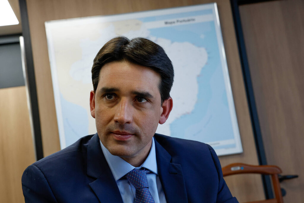 Um homem com cabelo castanho escuro e liso, vestindo um terno escuro e uma camisa clara, está sentado em um ambiente de escritório. Ele parece estar em uma conversa ou reflexão, olhando para a frente. Ao fundo, há um mapa do Brasil na parede, com destaque para a região da Amazônia. O ambiente é moderno, com paredes de madeira e uma cadeira de madeira visível ao lado.