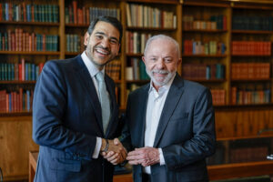 Dois homens de terno apertam as mãos em frente a estantes cheias de livros em uma biblioteca. Ambos sorriem e estão em ambiente interno com móveis de madeira.