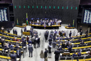 A imagem mostra o plenário da Câmara dos Deputados do Brasil, com várias pessoas em movimento. Há uma mesa central onde alguns deputados estão reunidos, e ao fundo, as bandeiras do Brasil e do Congresso. As cadeiras estão ocupadas por outros deputados, e as telas de votação estão visíveis nas laterais. A imagem tem um efeito de desfoque, sugerindo movimento.