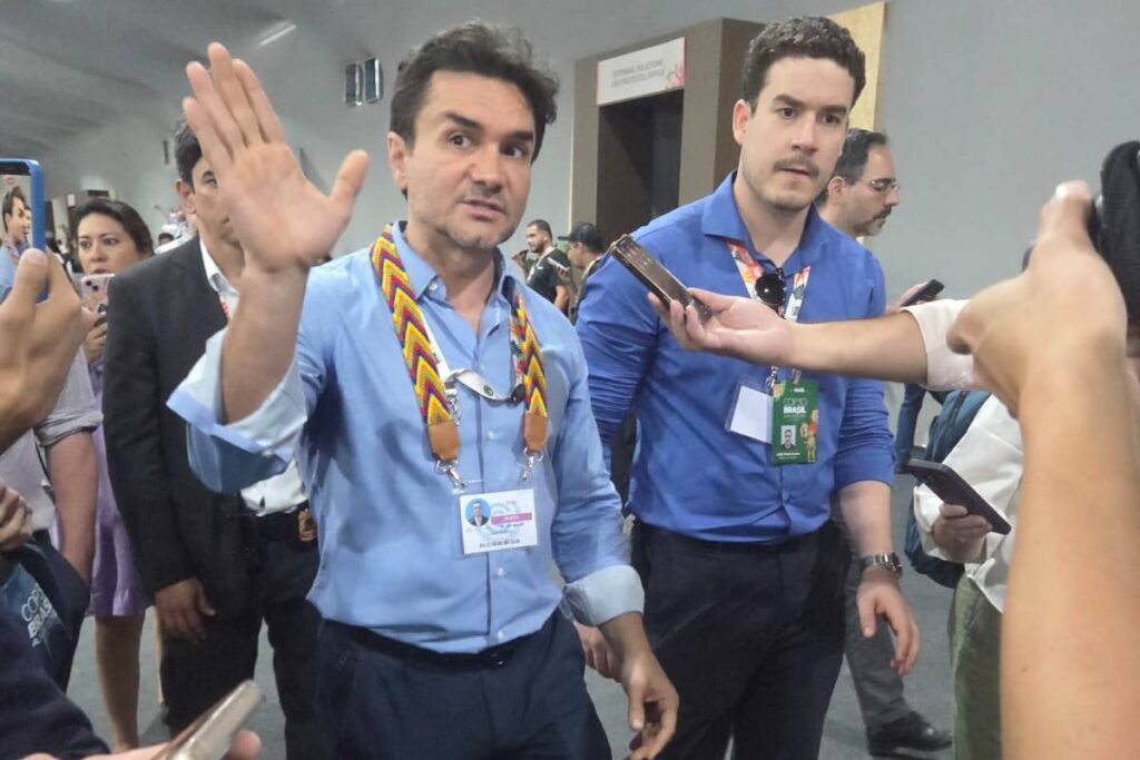 Dois homens de camisa azul e calça escura caminham em corredor interno. Um deles levanta a mão direita em gesto de cumprimento ou bloqueio, enquanto o outro observa. Pessoas ao redor seguram celulares, possivelmente para fotos ou vídeos.