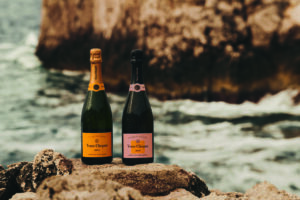 Duas garrafas de champanhe Veuve Clicquot, uma com rótulo amarelo e outra com rótulo rosa, estão posicionadas sobre pedras irregulares próximas ao mar. Ao fundo, ondas quebram contra uma formação rochosa grande e desfocada.