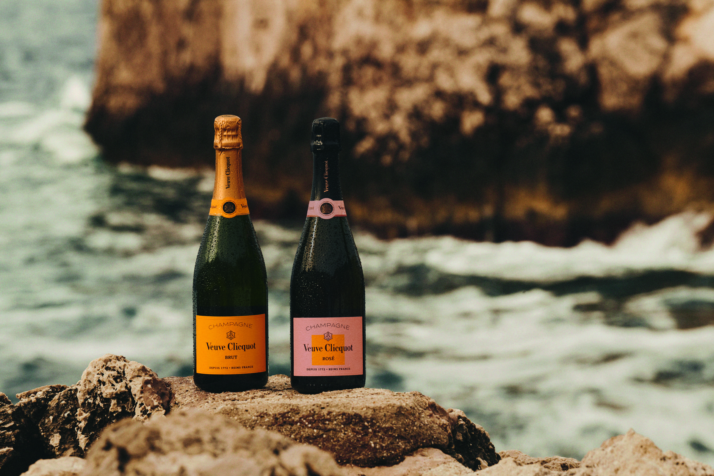 Duas garrafas de champanhe Veuve Clicquot, uma com rótulo amarelo e outra com rótulo rosa, estão posicionadas sobre pedras irregulares próximas ao mar. Ao fundo, ondas quebram contra uma formação rochosa grande e desfocada.