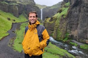 Homem sorridente veste jaqueta amarela e cinza, posando em trilha de terra ao lado de rio estreito. Ao fundo, cachoeira desce entre penhascos verdes e rochosos sob céu nublado.