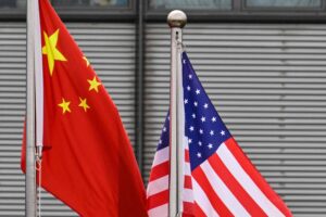 Bandeira da China à esquerda e bandeira dos Estados Unidos à direita, ambas hasteadas em postes metálicos diante de fachada com janelas e persianas fechadas.