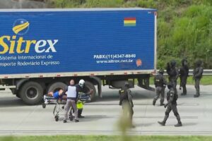 Equipe de resgate empurra maca com pessoa deitada na pista ao lado de caminhão azul da Sitrex. Policiais à direita caminham pela rodovia, com vegetação ao fundo.