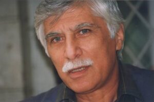 Homem idoso com cabelo grisalho e bigode branco olha diretamente para a câmera. Fundo desfocado com padrão geométrico.