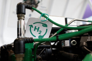 Detalhe de conexões e tubos verdes em equipamento industrial com etiqueta mostrando símbolo de H2 e seta circular indicando ciclo.