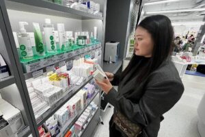 Mulher de cabelo longo e preto, vestindo casaco cinza escuro, segura e examina embalagem de produto em prateleira de farmácia. Prateleiras exibem diversos cosméticos e medicamentos organizados por categorias.