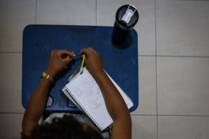 A imagem mostra uma mesa azul com uma pessoa escrevendo em um caderno. A pessoa está segurando um lápis amarelo com a mão direita. O caderno está aberto e possui anotações. Ao lado, há um copo preto com tampa. O ambiente parece ser interno, com piso claro.