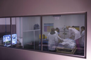 Quatro profissionais de saúde com jalecos brancos atendem paciente deitado em maca dentro de sala hospitalar. Equipamentos médicos e monitores estão visíveis ao fundo e à esquerda.