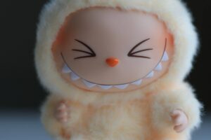 A imagem mostra um boneco de pelúcia com um sorriso largo. O boneco é de cor laranja claro e está vestido com um traje que parece uma fantasia de animal, com orelhas e dentes visíveis. O fundo é desfocado, destacando o boneco.
