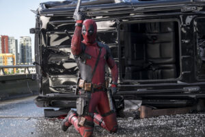 deadpool-filme-heroi-ajoelhado-em-cena-de-acao-com-veiculo-destruido-ao-fundo