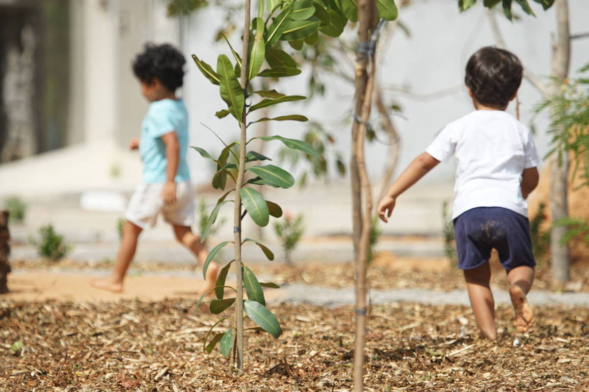 A imagem mostra duas crianças pequenas brincando ao ar livre em um ambiente natural. Uma criança está vestida com uma camiseta azul e shorts claros, enquanto a outra usa uma camiseta branca e shorts escuros. Elas estão em um espaço com solo coberto de folhas secas e algumas plantas ao redor. A criança à esquerda está de costas, enquanto a da direita está se afastando.