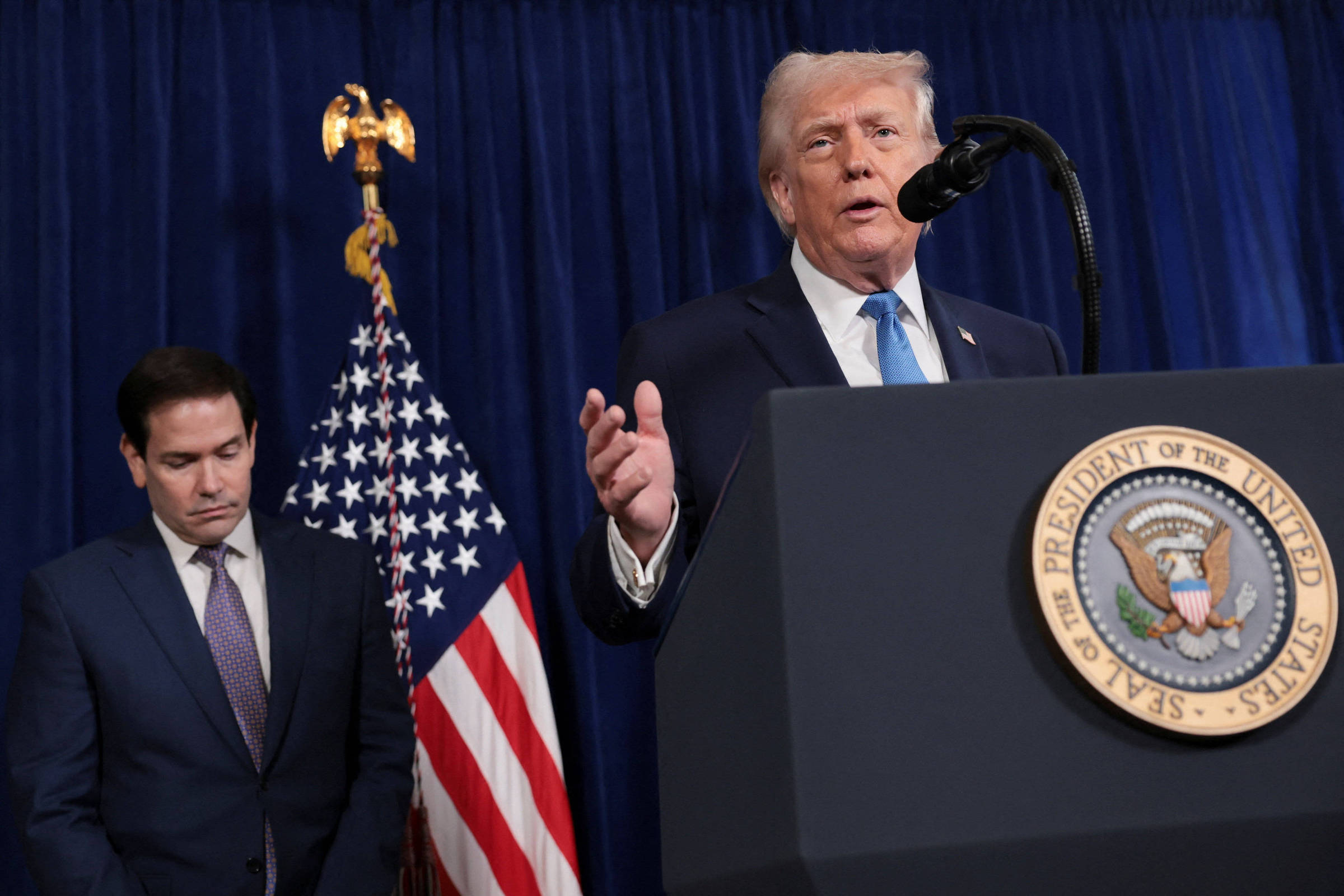 Ex-presidente dos Estados Unidos fala em púlpito com selo presidencial, enquanto senador está ao lado com olhar para baixo. Bandeira americana e cortina azul ao fundo.