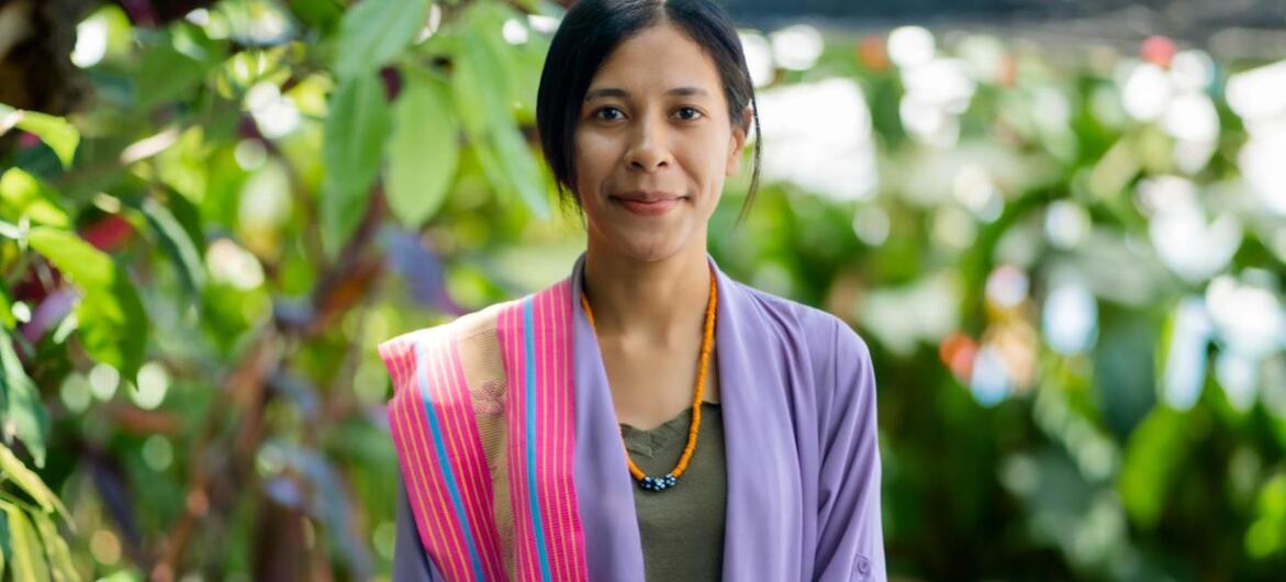 Um retrato de Joanita Pinto, uma mulher de cabelo escuro, usando um casaco roxo e um colar de laranja, sorrindo diante de folhas verdes exuberantes.