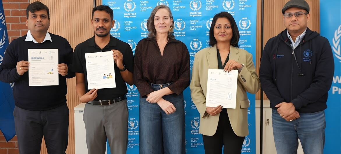 Cinco indivíduos estão de pé em frente a um cenário do Programa Mundial de Alimentos, segurando certificados, celebrando como vencedores do hackathon.