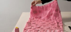 Uma pessoa segura um pedaço de tecido rosa com padrões de textura circular, com uma bobina de fio rosa e uma fita de medição amarela em uma mesa branca.
