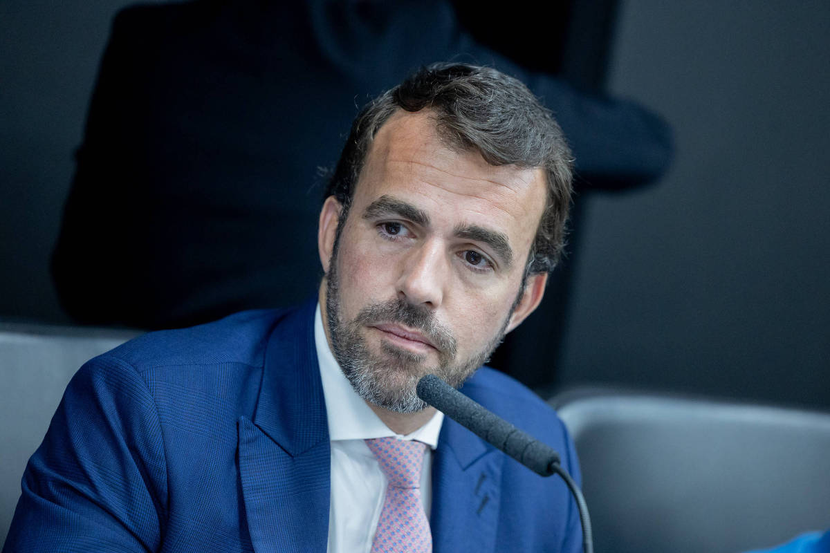 Homem de cabelos curtos e barba aparada veste terno azul, camisa branca e gravata rosa, sentado diante de microfone em ambiente formal.