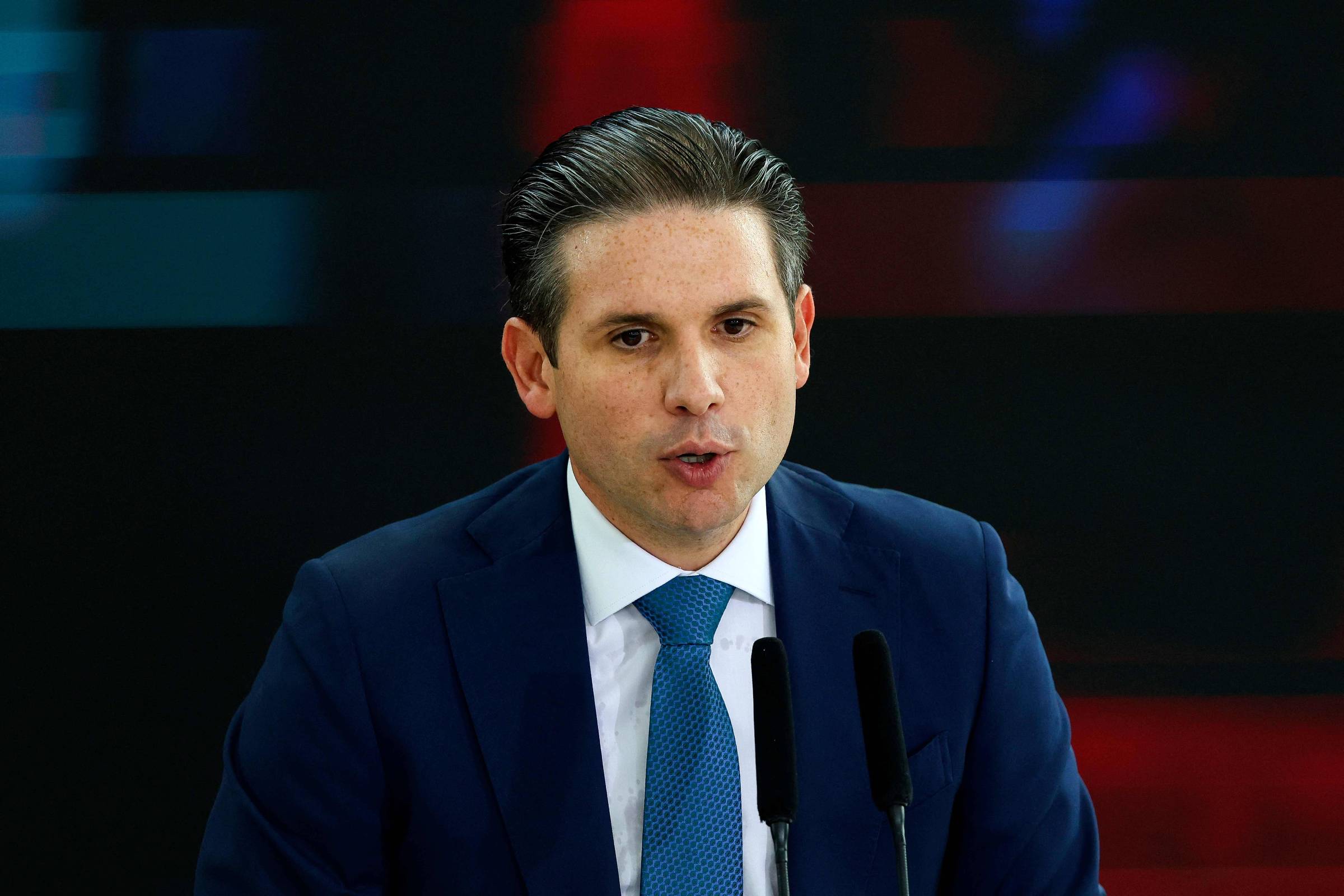 Homem de cabelo escuro penteado para trás veste terno azul escuro, camisa branca e gravata azul, falando em dois microfones diante de fundo escuro com luzes desfocadas vermelhas e azuis.