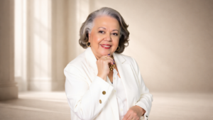 Myrinha Vasconcellos em retrato institucional, vestindo blazer branco, representando o conceito de longevidade ativa e protagonismo feminino.