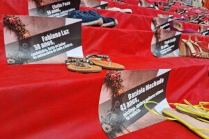 Várias sandálias femininas dispostas sobre degraus cobertos por tecido vermelho, cada par acompanhado por uma placa com nome, idade e causa da morte de mulheres vítimas de violência.